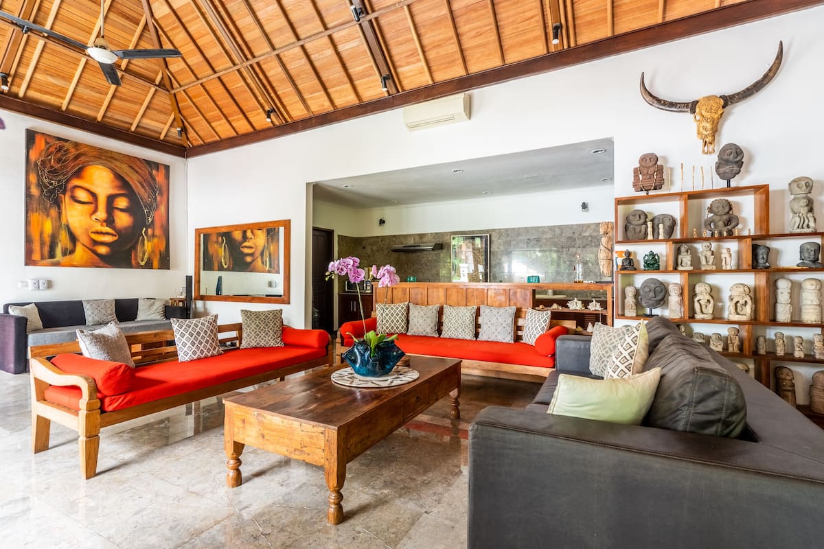A spacious living room with a high ceiling, wooden beams, a red sofa set, a coffee table, and a collection of sculptures on the wall.