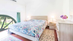 Quarto com cama, mesa de cabeceira, luminária, cômoda e janela com cortinas.