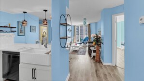 A modern kitchen with white cabinets, a dishwasher, and a sink. There is a dining area with a table and chairs, and a living room with a sofa and a coffee table. - Oceanfront condo with ocean views! (North Topsail Beach)