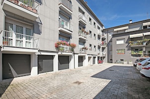A modern building with multiple balconies, some with flower boxes, and a paved driveway with parked cars.