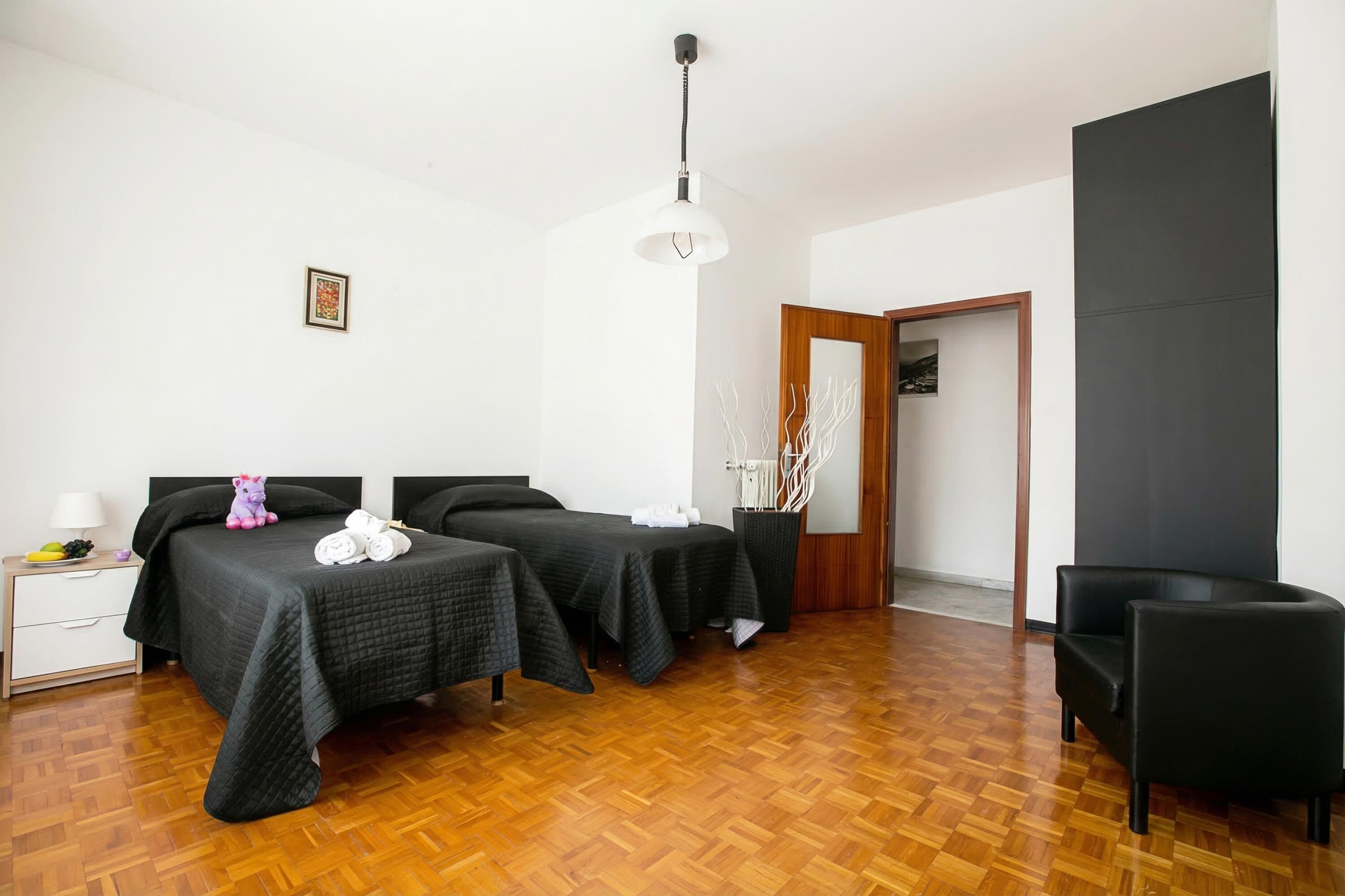 A hotel room with two beds, a wooden floor, a black chair, a bedside table with a lamp, and a framed picture on the wall.