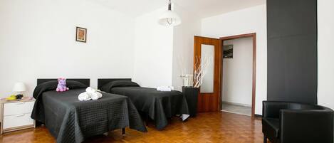 A hotel room with two beds, a wooden floor, a black chair, a bedside table with a lamp, and a framed picture on the wall.