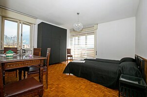 A bedroom with a wooden floor, a bed, a desk with a chair, a window with blinds, and a hanging light fixture.