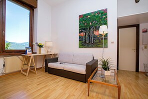 A living room with a sofa, a table, a lamp, a painting, and a view through a window. - Appartamento Villa Lina (Riva del Garda)