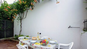 A patio table set with plates, cutlery, and a fruit bowl, surrounded by white chairs.
