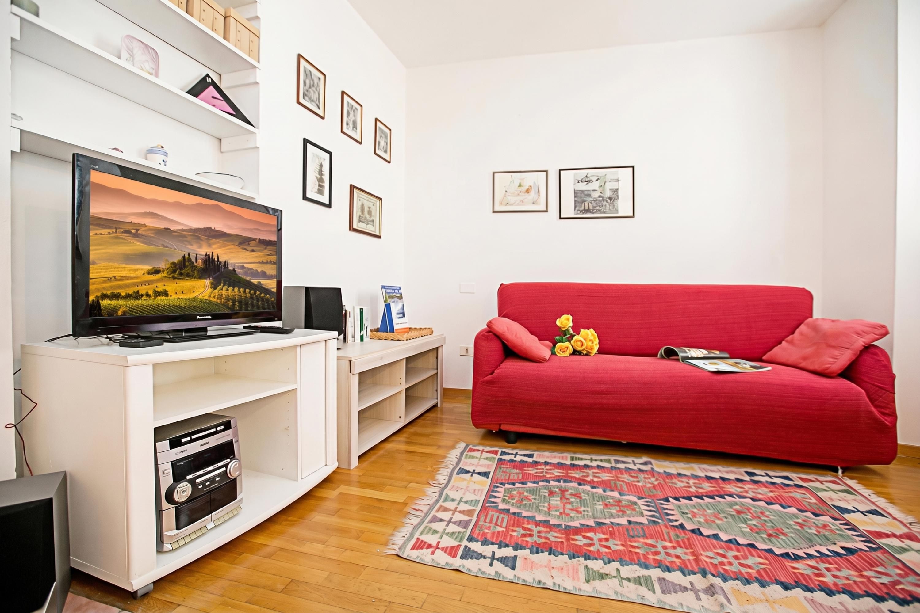 Un salon doté d’un téléviseur à écran plat, d’un canapé rouge, d’un tapis à motifs et d’un meuble de rangement blanc avec des étagères et un placard.