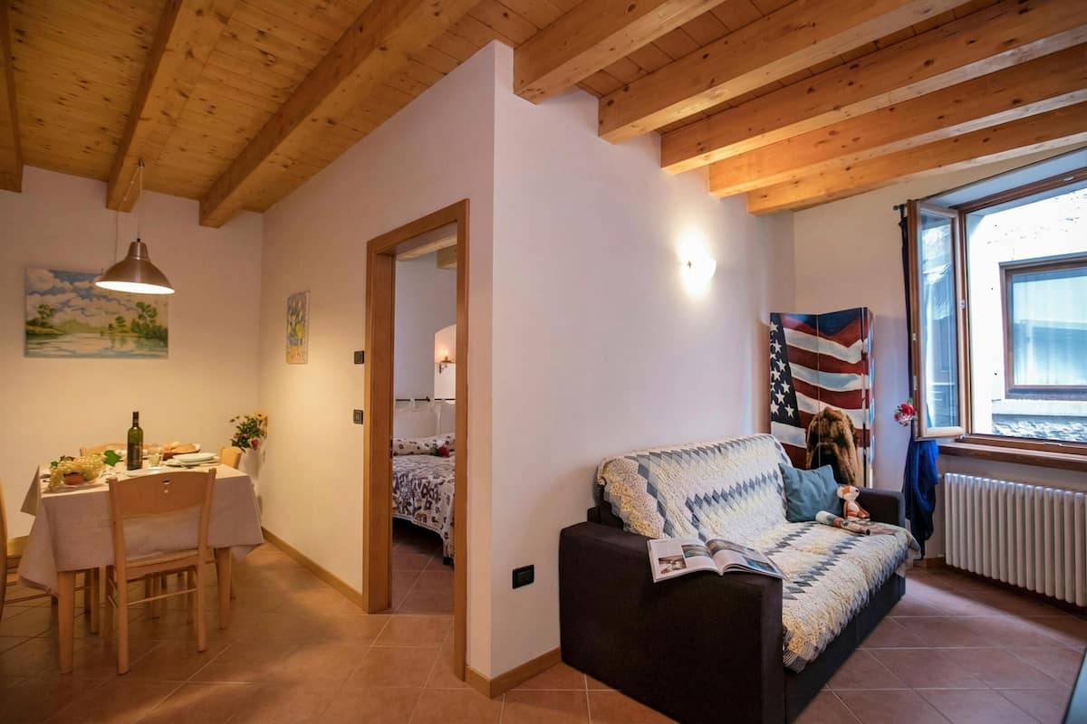 A cozy living room with a wooden ceiling, a dining area with a table and chairs, a sofa with a quilt, and a window with an American flag.