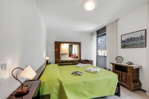 A hotel room with a green quilted bedspread, a bedside lamp, a mirror, a wooden dresser, and a framed picture on the wall. - Appartamento Amy (Riva del Garda)