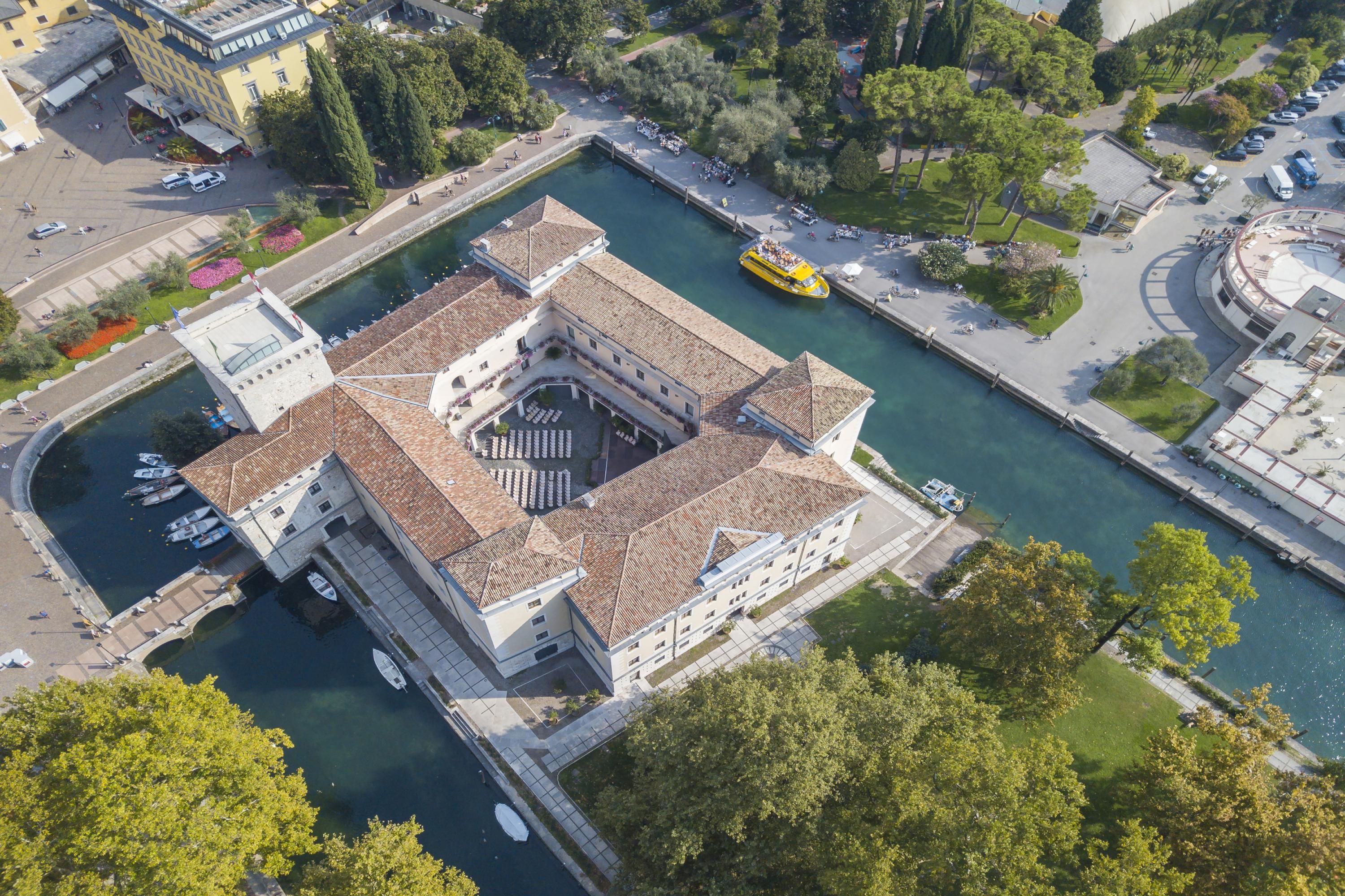 An aerial view of a historic building with a central courtyard, adjacent to a canal with boats and a yellow boat docked.
