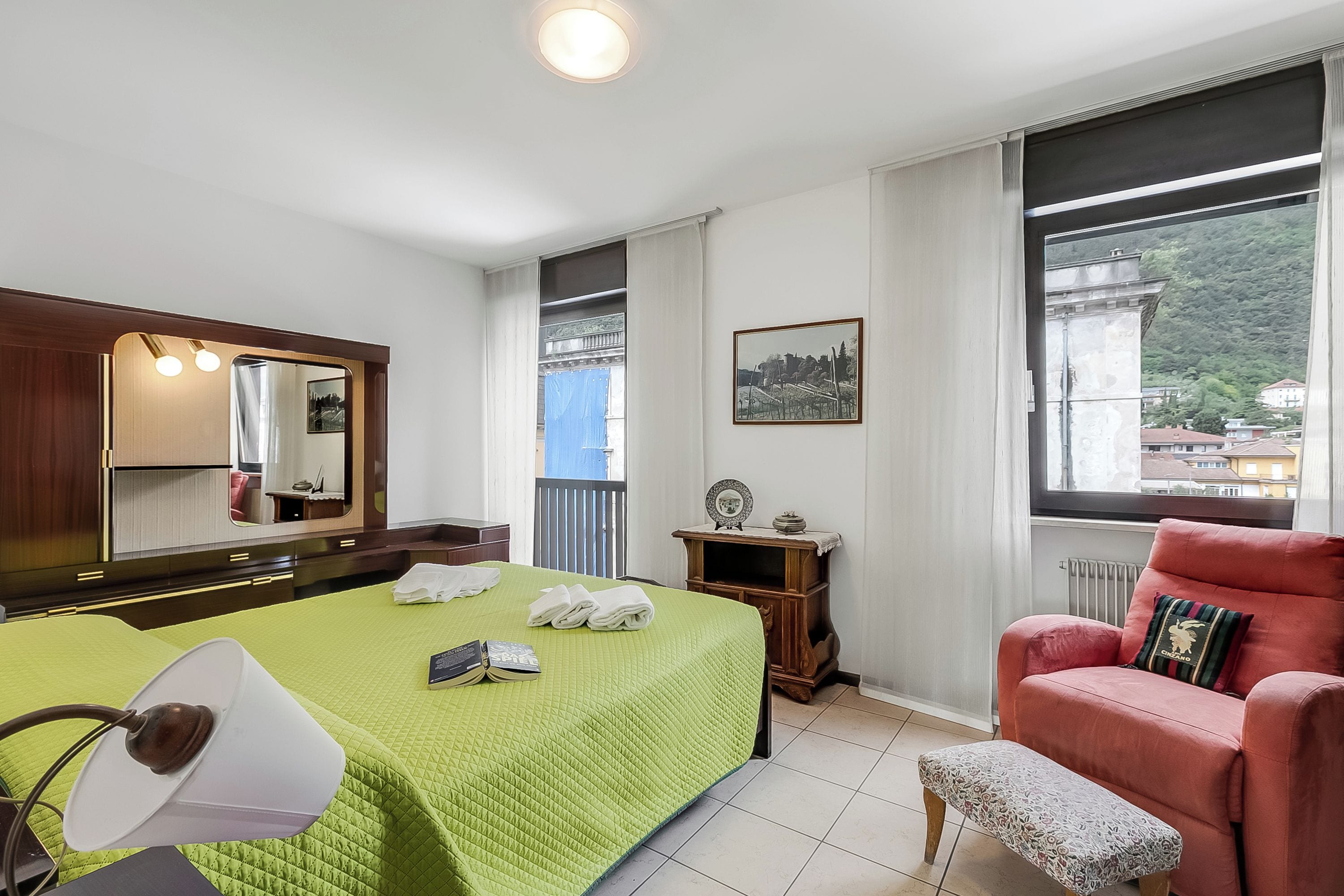A hotel room with a bed, a chair, a small table, a mirror, and a window with a view of buildings and hills.