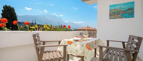 A balcony with two wooden chairs, a table with a floral tablecloth, and a painting on the wall.