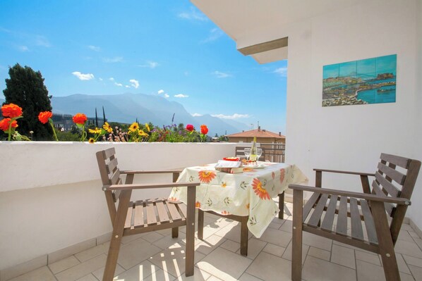 A balcony with two wooden chairs, a table with a floral tablecloth, and a painting on the wall.