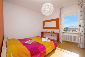 A bedroom with a bed, a bedside table, a mirror, a window with curtains, and a hanging light fixture.