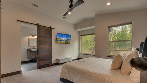 A bedroom with a large bed, a sliding door to a bathroom, a ceiling fan, and a large window with a view of trees. - AC, Large Upright Arcade, Dreamy Kitchen, Designer Furnished, Grill, Fast Wifi, Spa – The Glenmore (South Lake Tahoe)