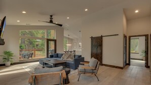 A spacious living room with a large sectional sofa, a coffee table, and a fireplace. There is a sliding barn door and a view of the outdoors through large windows. - AC, Large Upright Arcade, Dreamy Kitchen, Designer Furnished, Grill, Fast Wifi, Spa – The Glenmore (South Lake Tahoe)
