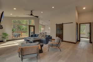 A spacious living room with a large sectional sofa, a coffee table, and a fireplace. There is a sliding barn door and a view of the outdoors through large windows.