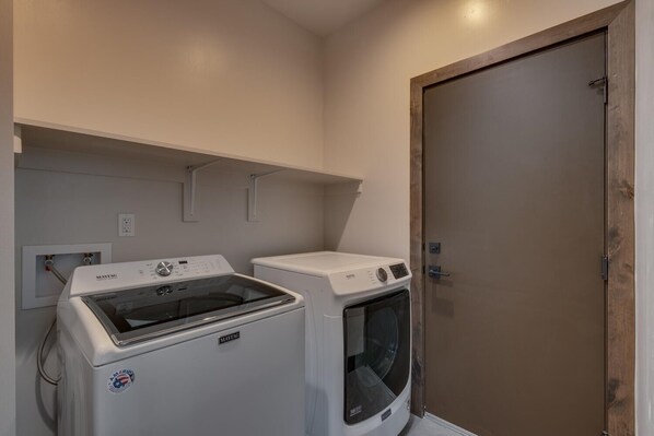 Een wasserij met een Maytag wasmachine en droger, een houten deur en een witte muur met een stopcontact.