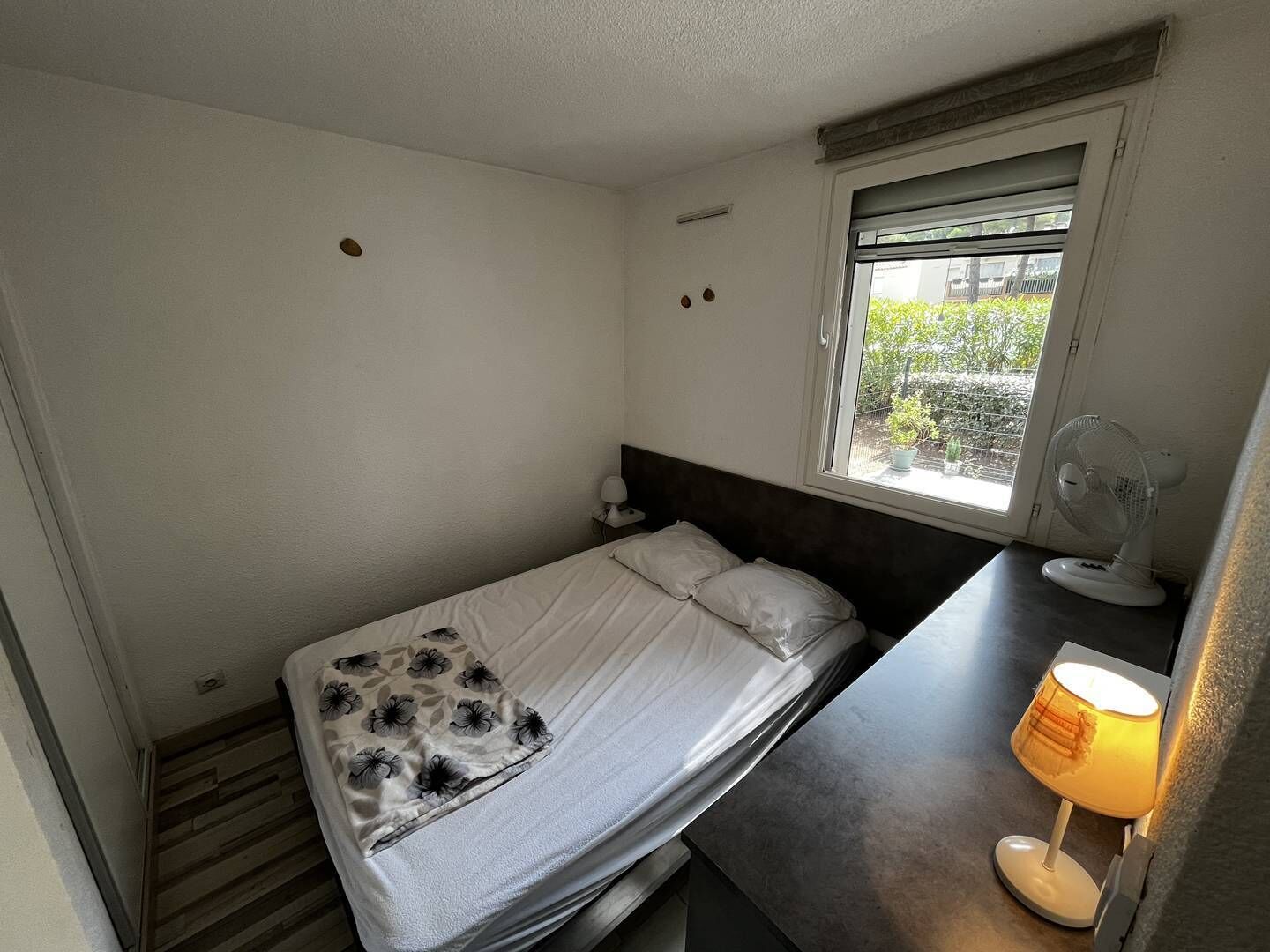 A small, single-bed room with a window, a bedside table with a lamp, and a wall-mounted fan.