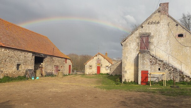 Ländliche Szenerie mit Steinhäusern, einem Feldweg und einem Regenbogen im Hintergrund.
