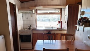 Eine kleine Küche mit Herd, Backofen und Spüle. Ein Esstisch mit Stühlen und ein Fenster mit Jalousien sind ebenfalls vorhanden.