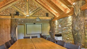 Ein Konferenzraum mit einem langen Holztisch, Korbstühlen, einer Projektorleinwand und einer Steinwand.