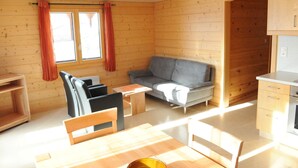 Ein gemütliches Blockhäuschen mit Holzvertäfelung, einem grauen Sofa, einem Esstisch mit Stühlen und einer Küchenzeile.