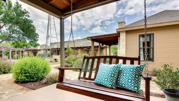 A wooden swing bench with cushions, a covered porch, and a building in the background.
