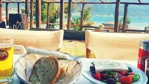 A table with a meal, a beer mug, and a Coca-Cola can, set against a scenic coastal backdrop. - Villa in Corfu Near Porto Timoni Beach (Corfu)