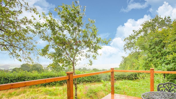 A wooden deck with a metal table and chairs, surrounded by a wooden railing and a view of greenery and hills.