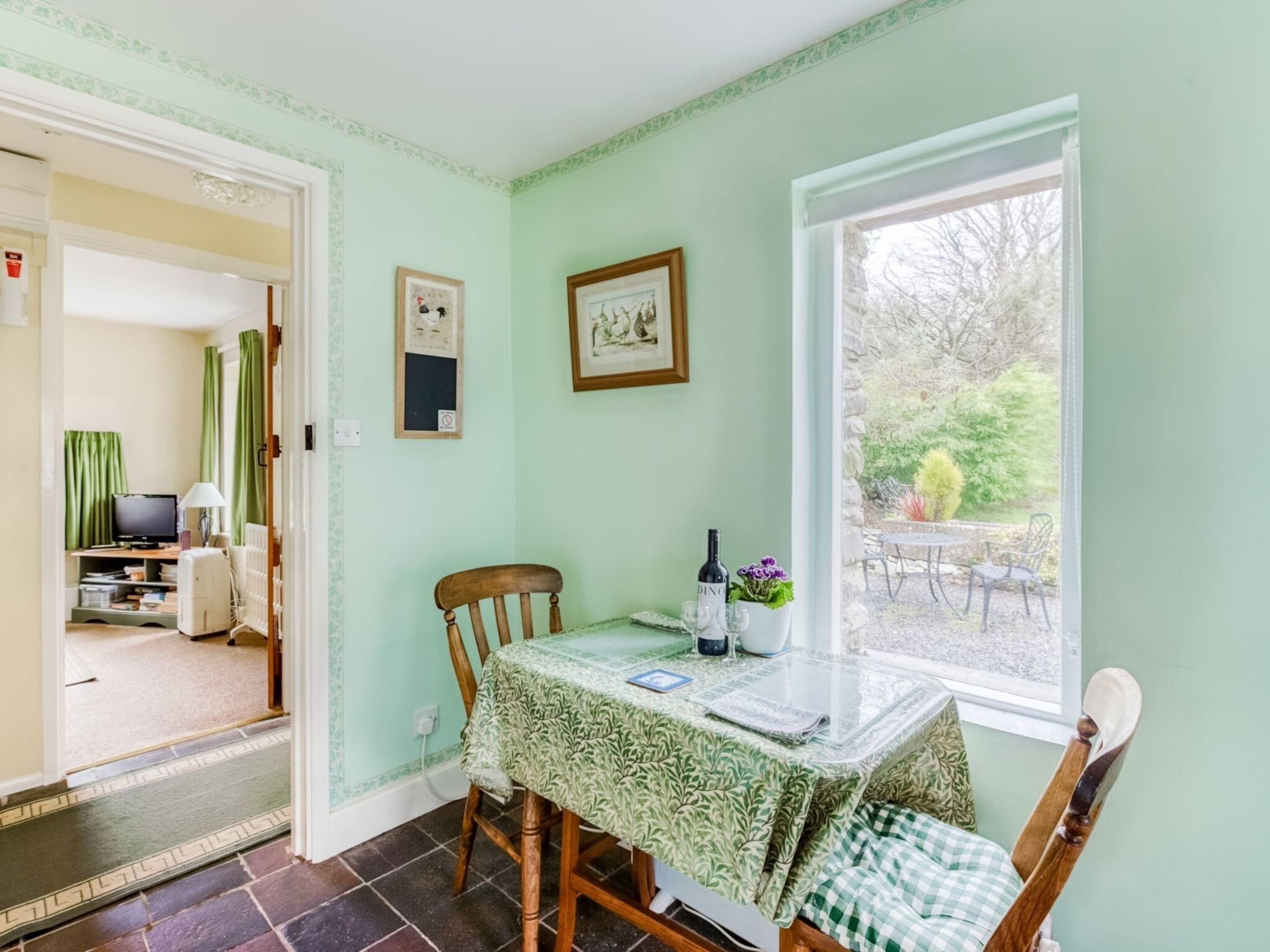 A dining area with a table set for two, a chair, a window with a view of the garden, and a door leading to another room.