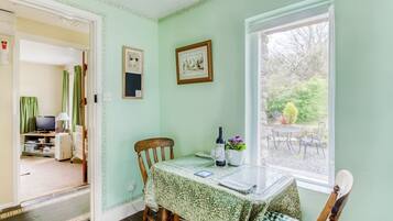 A dining area with a table set for two, a chair, a window with a view of the garden, and a door leading to another room.