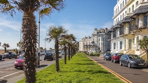 Une rue bordée de palmiers et de voitures garées, qui mène à une série de bâtiments de style européen traditionnel, dotés de balcons.