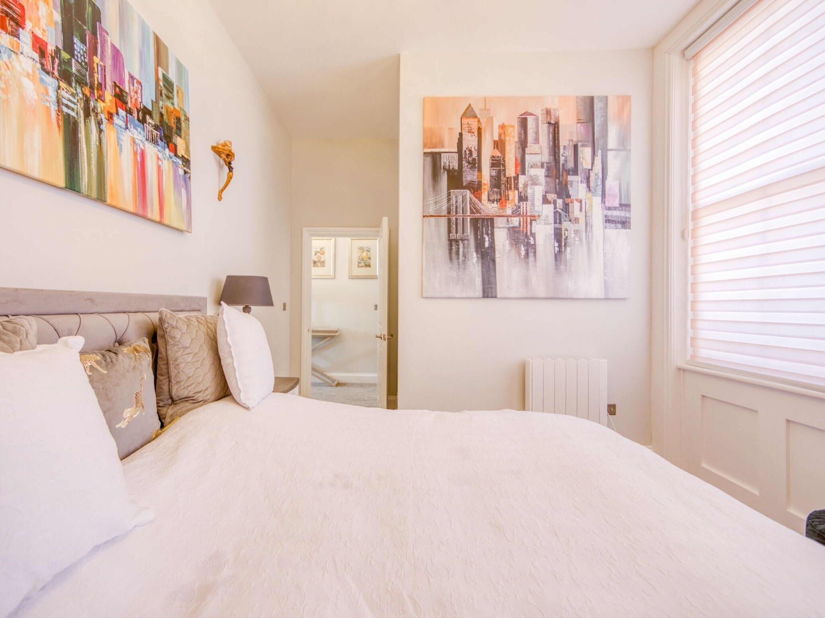A bedroom with a large bed, a cityscape painting, and a view of a city skyline through the window.