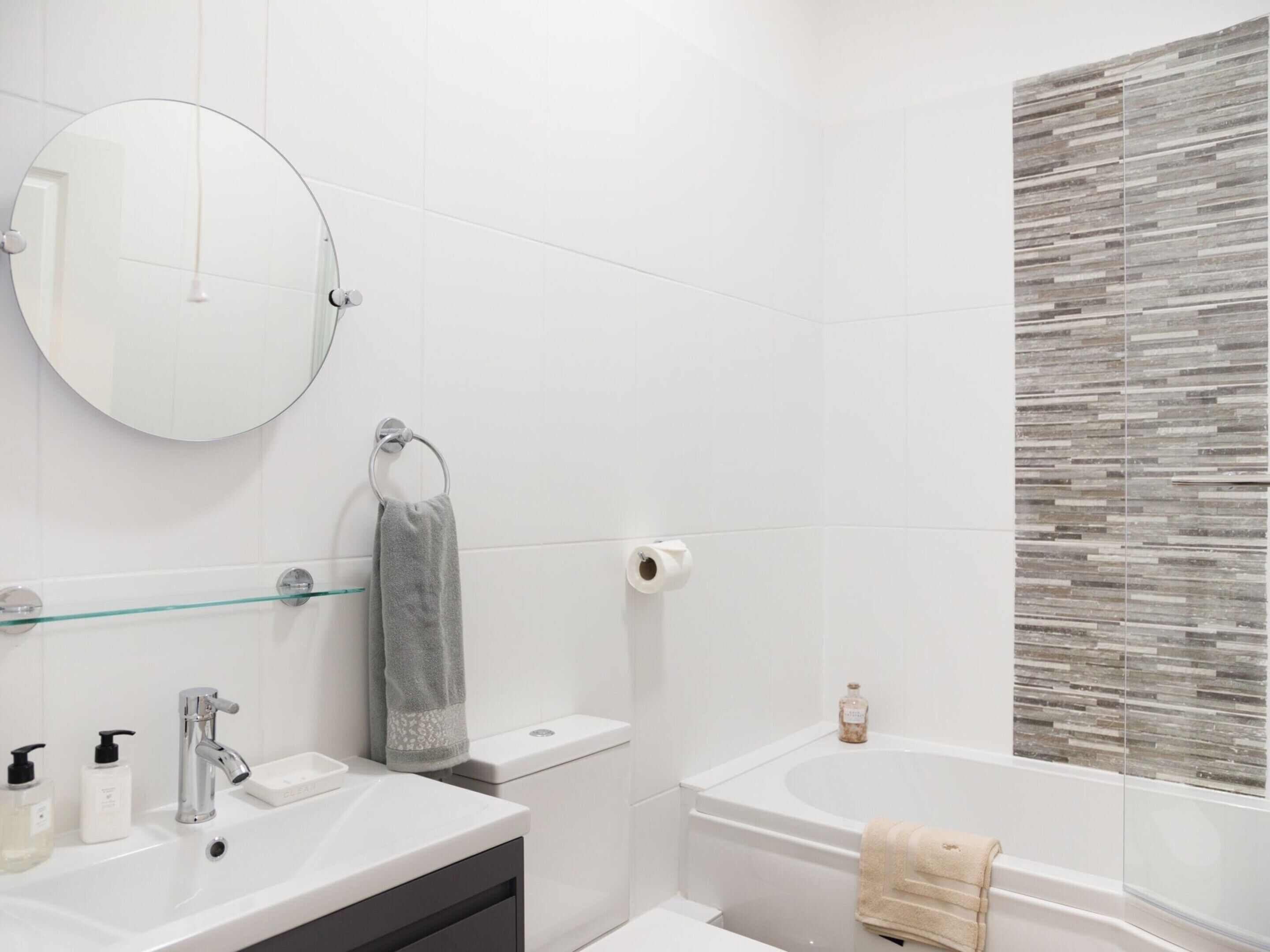 A modern bathroom with a round mirror, a toilet, a sink with soap dispensers, and a bathtub.
