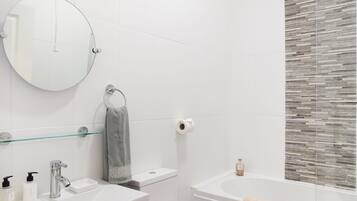 A modern bathroom with a round mirror, a toilet, a sink with soap dispensers, and a bathtub.
