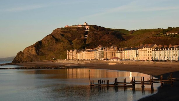 A coastal town with historic buildings, a prominent hill with a structure on top, and a calm waterfront. - Holiday Home in Ceredigion Near Seashore (Llanon)