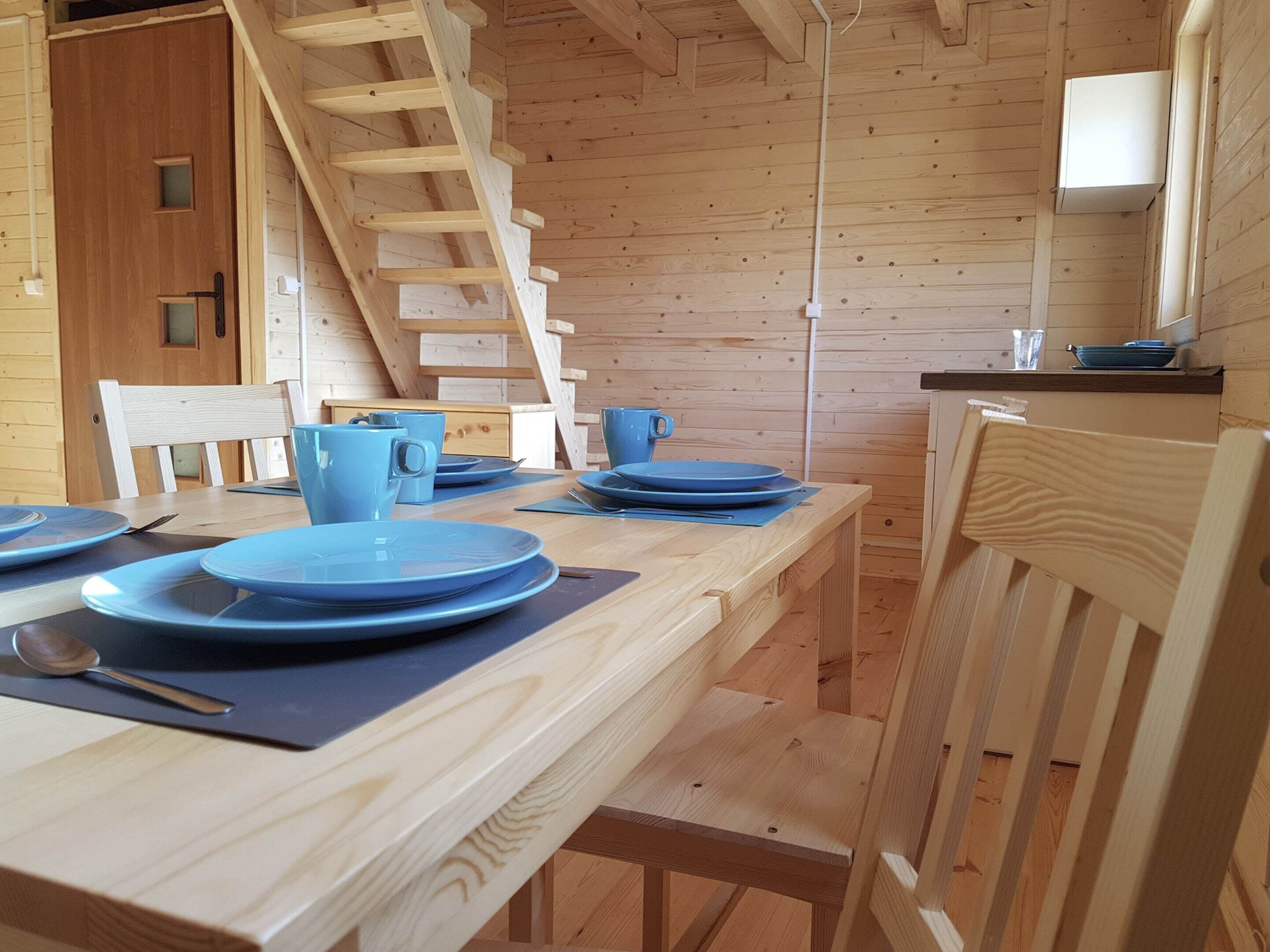 A wooden interior with a dining table set for four, a wooden staircase, and a kitchen area with a sink and countertop.