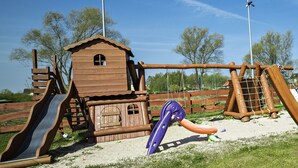 Een houten speelconstructie met een glijbaan en klimelementen, gelegen in een grasrijk gebied met bomen en lantaarns op de achtergrond.
