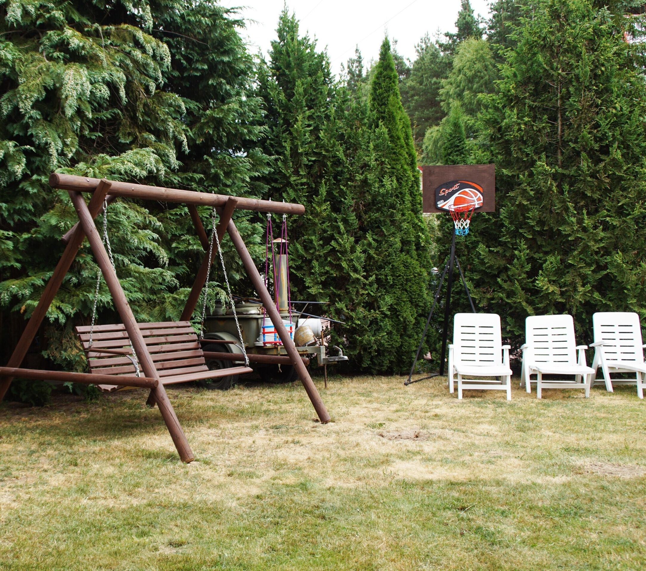 Altalena di legno, canestro da basket e tre sedie da giardino bianche in un cortile circondato da alberi.