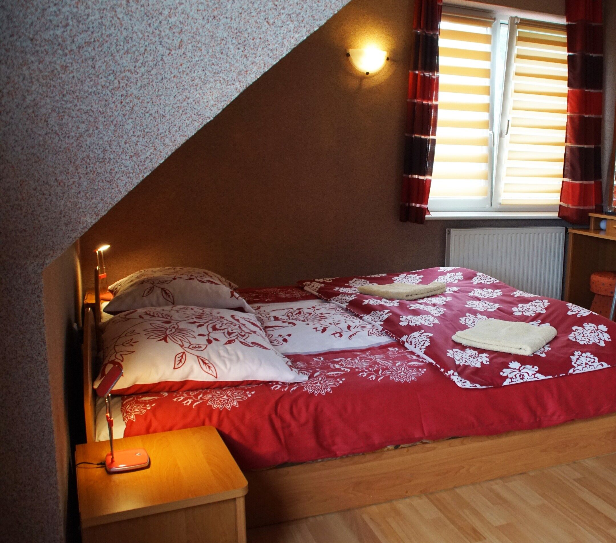 A bedroom with a sloped ceiling, a bed with a red and white floral bedspread, two pillows, a bedside lamp, and a window with blinds.
