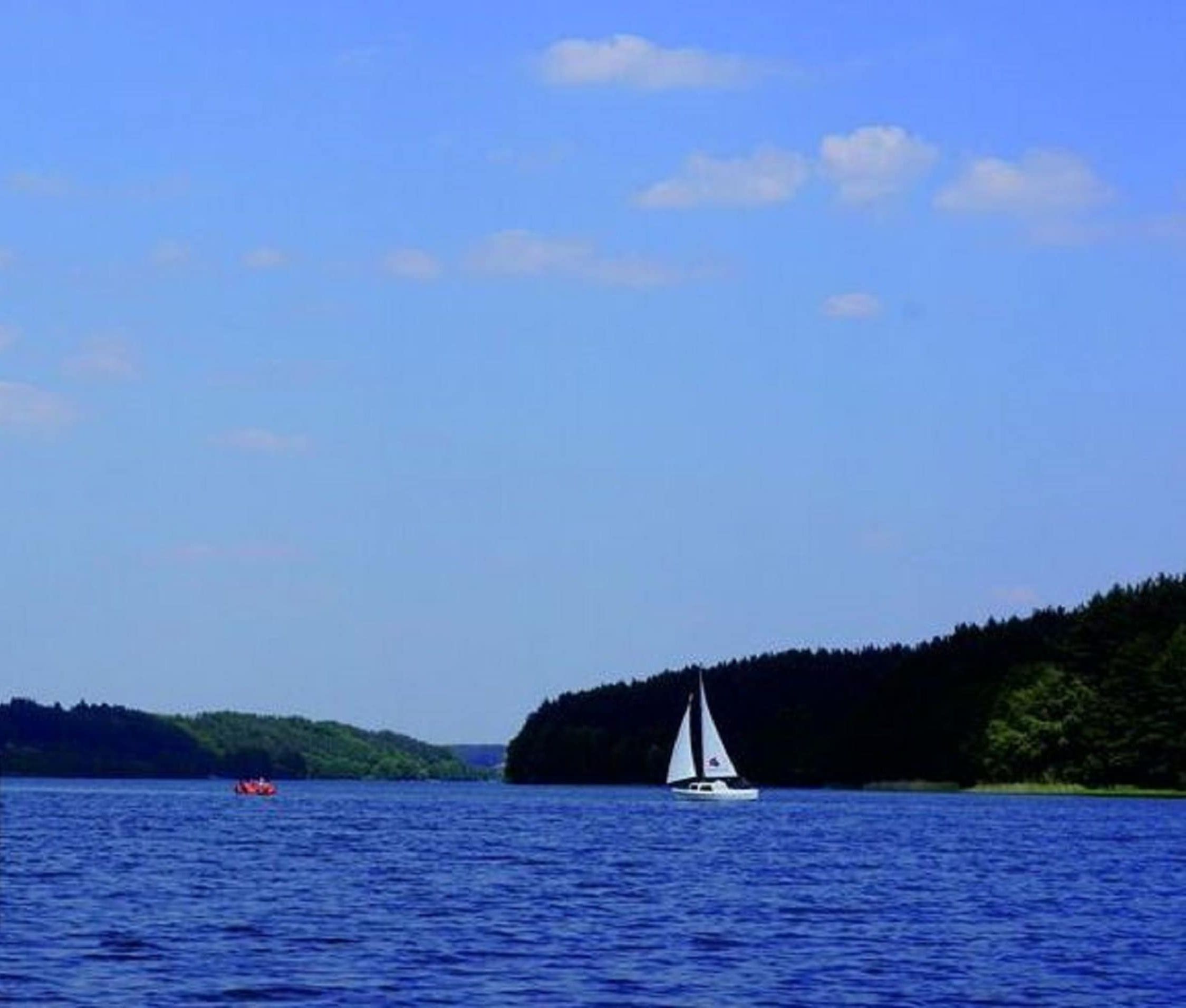 Un lac paisible, avec un voilier et un bateau rouge au loin, le tout entouré de terres boisées.