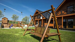 A wooden swing set and a hammock in a grassy area with a wooden building in the background. - Lovely House Overlooking the Lake, Near the Sea, Ideal for 9 People (Mielno)
