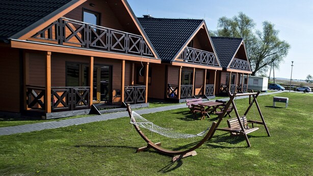 A row of wooden cabins with balconies, a hammock, and picnic tables on a grassy lawn. - Lovely House Overlooking the Lake, Near the Sea, Ideal for 9 People (Mielno)