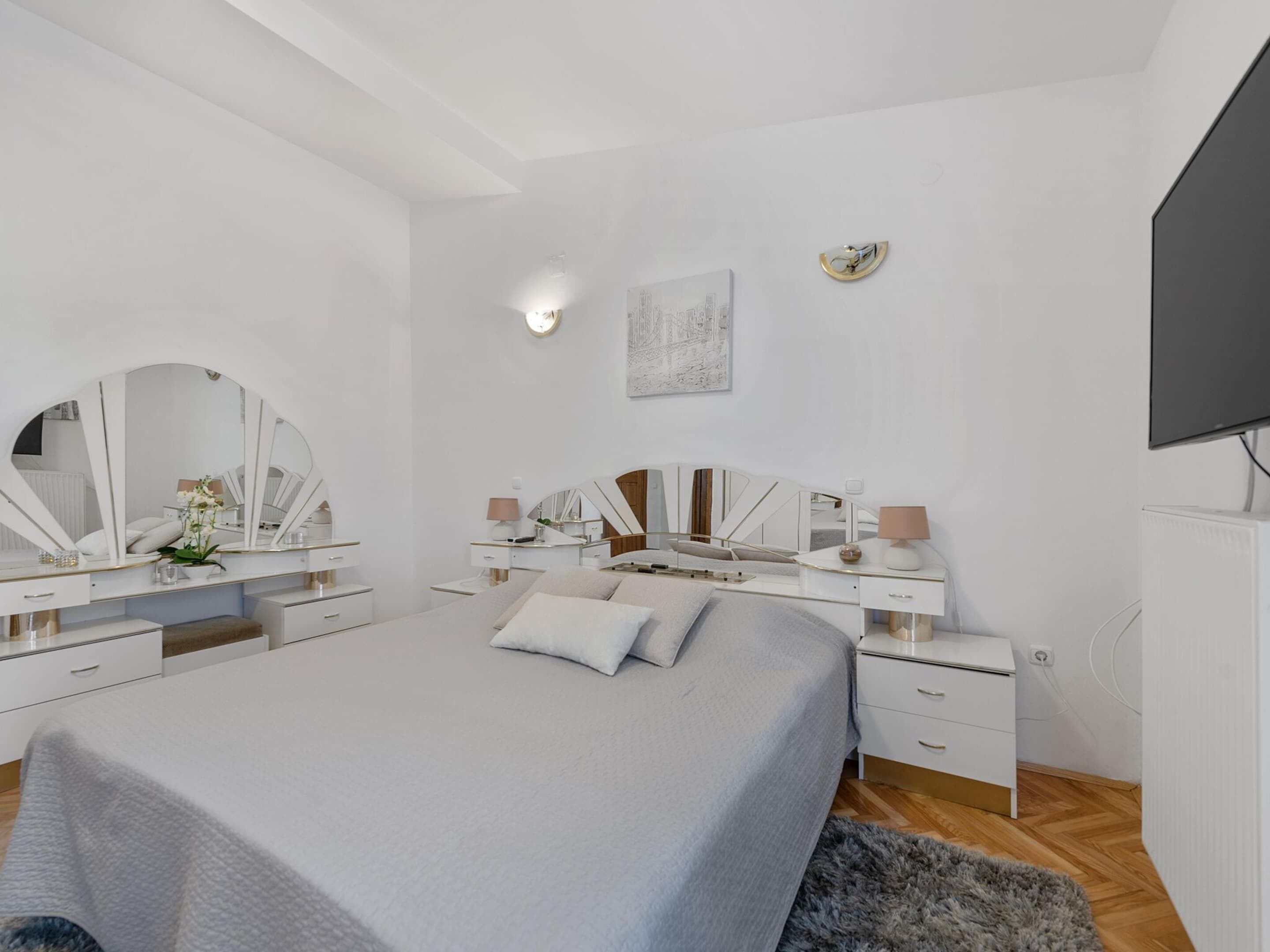 A modern bedroom with a large bed, two bedside tables with mirrors, a flat-screen TV, and a small desk.
