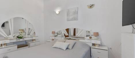 A modern bedroom with a large bed, two bedside tables with mirrors, a flat-screen TV, and a small desk.