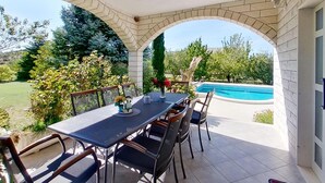 Un porxo cobert amb taula i cadires, una piscina i vista a un jardí ben cuidat.