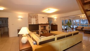 Un salón amplio con sofá, televisor y chimenea. Se ve una cocina al fondo.