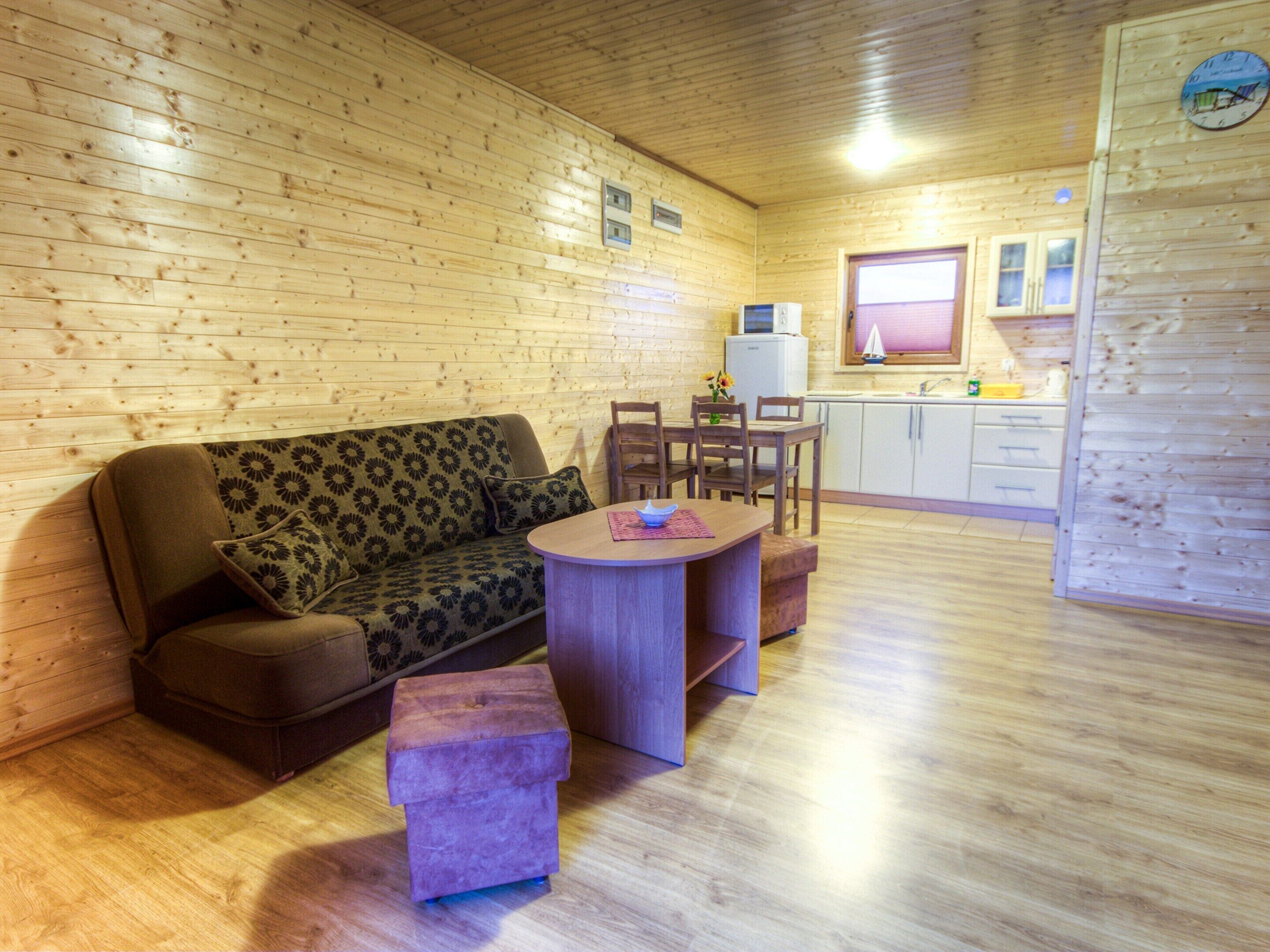 A cozy cabin interior with a wooden floor, a sofa with a floral pattern, a small round table, and a kitchenette area.