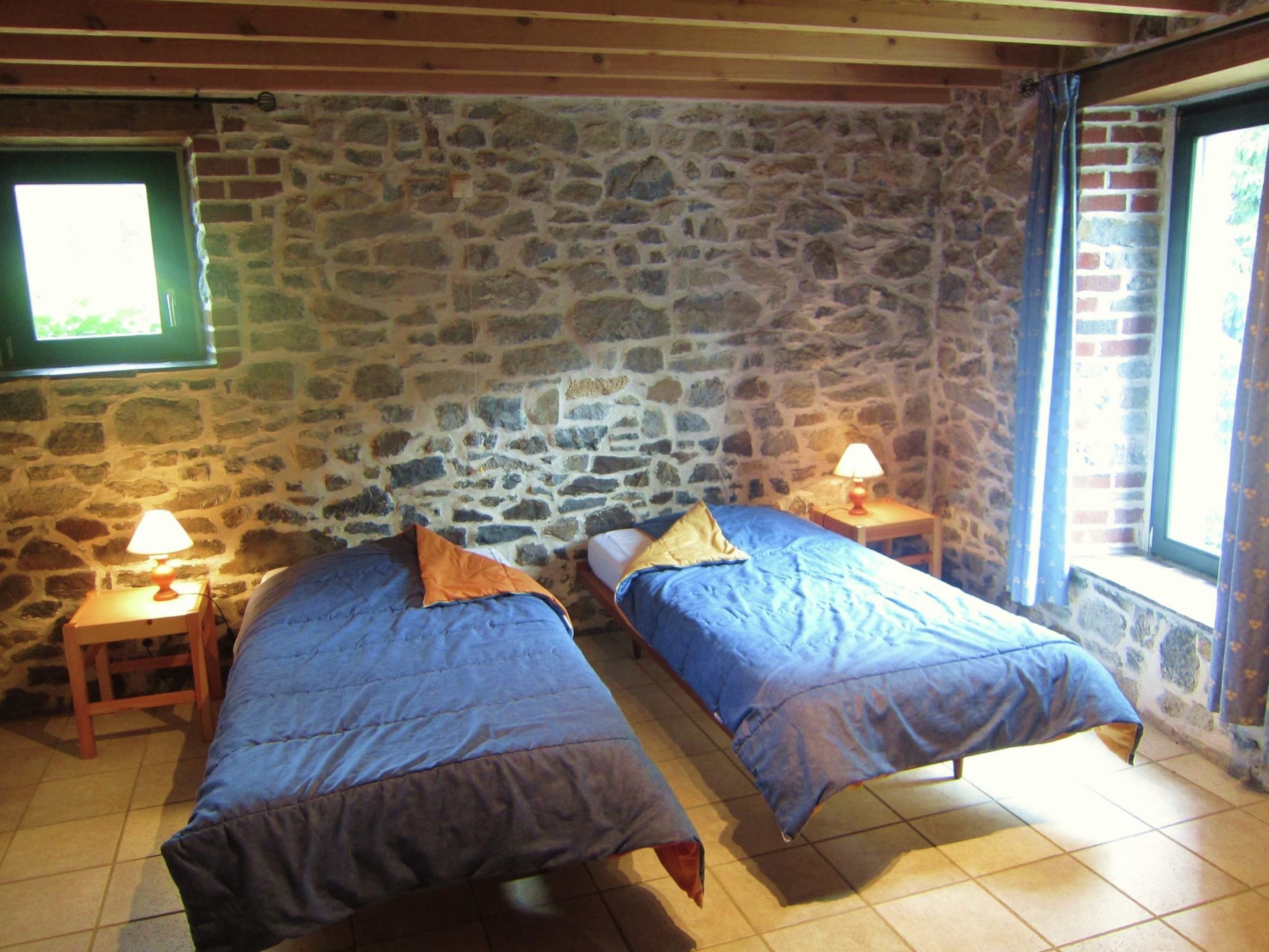 A room with two beds, stone walls, a window, and a lamp on a nightstand.