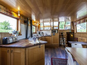 A wooden cabin-style kitchen with a wooden countertop, a toaster, a kettle, a microwave, and a sink. There are windows with curtains, a wooden ceiling, and a view of greenery outside. - Boat Getaway Near Lake (Houthulst)